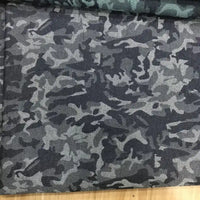 Le tissu denim jacquard camouflage extensible au mètre de Longon Craft France est présenté sur une surface en bois, avec un autre tissu denim camouflage coloré partiellement visible au-dessus.