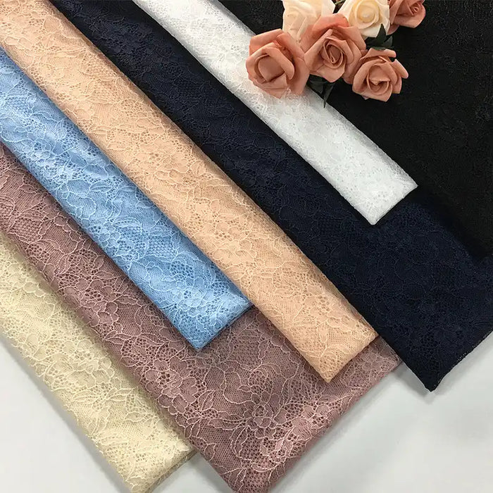 Sept pièces pliées de tissu dentelle extensible multicolore de Longan Craft France sont présentées à côté de roses artificielles de couleur pêche. Elles sont parfaites pour coudre de la lingerie, des vêtements ou des jupes avec notre "Tissu dentelle extensible au mètre" de haute qualité.