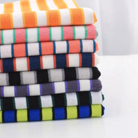 Une pile bien rangée de tissu piqué de coton rayé au mètre de Longan Craft France - un tissu de coton extensible tricolore pour les t-shirts DIY - présente des rayures horizontales vibrantes d'orange, de rose, de noir, de bleu et de vert sur une surface blanche.
