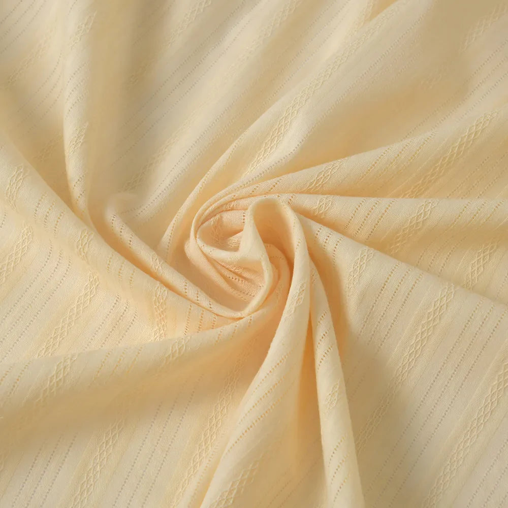 Le Tissu dobby jacquard ajouré rayé au mètre de Longan Craft France, un tissu en pur coton idéal pour l'habillement, est présenté en jaune clair avec un motif subtil de losanges et de rayures, tourbillonné à plat pour montrer sa texture douce et ses plis élégants.