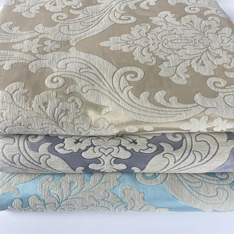 Trois tissus pliés de Longan Craft France, présentant des motifs jacquard vintage damassés à fleurs et à feuilles en chenille beige, grise et bleu clair, évoquent l'élégance classique - idéale comme tissu d'ameublement épais vendu au mètre.