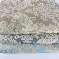 Trois tissus pliés de Longan Craft France, présentant des motifs jacquard vintage damassés à fleurs et à feuilles en chenille beige, grise et bleu clair, évoquent l'élégance classique - idéale comme tissu d'ameublement épais vendu au mètre.
