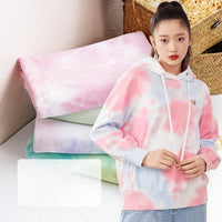 Une jeune femme porte un sweat à capuche rose et bleu en Tissu French Terry en coton tie-dye au mètre de Longan Craft France, avec un cordon de serrage blanc et un écusson en forme de papillon. Elle se tient devant des sweats à capuche pastel ; un panier tressé se trouve à l'arrière-plan.