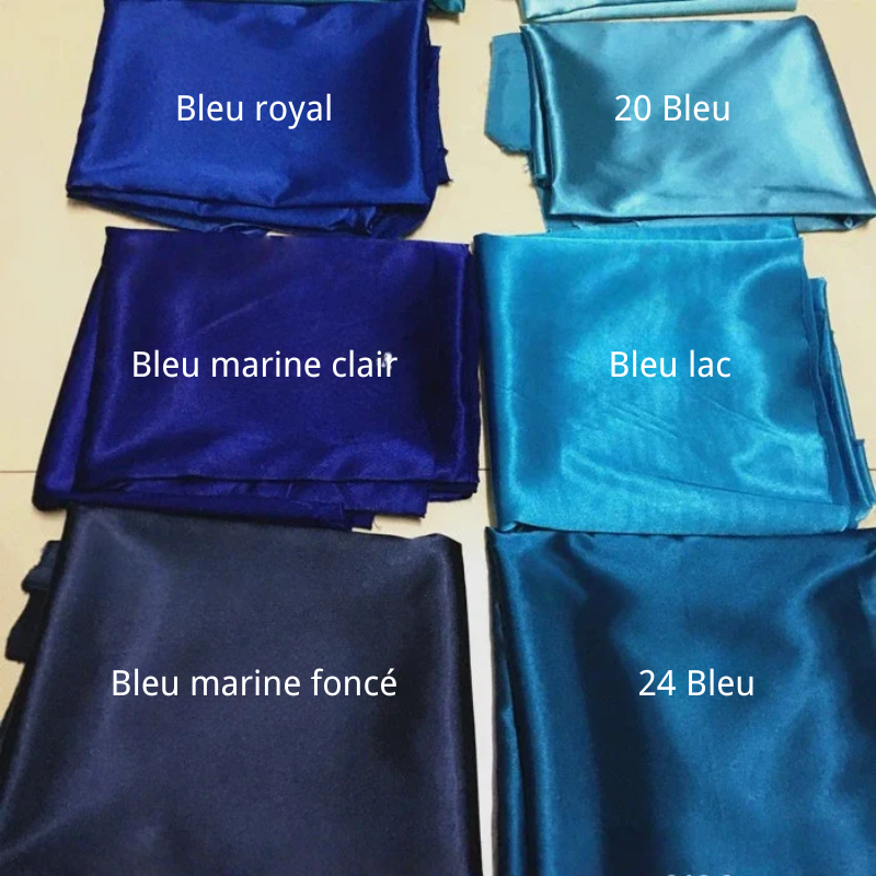 Six pièces pliées de tissu satin polyester uni au mètre de Longan Craft France dans différentes nuances de bleu, chacune étiquetée avec un nom de couleur français comme "Bleu royal" et "Bleu marine foncé", sont présentées sur une surface jaune, idéale pour les vêtements ajustés et confortables.