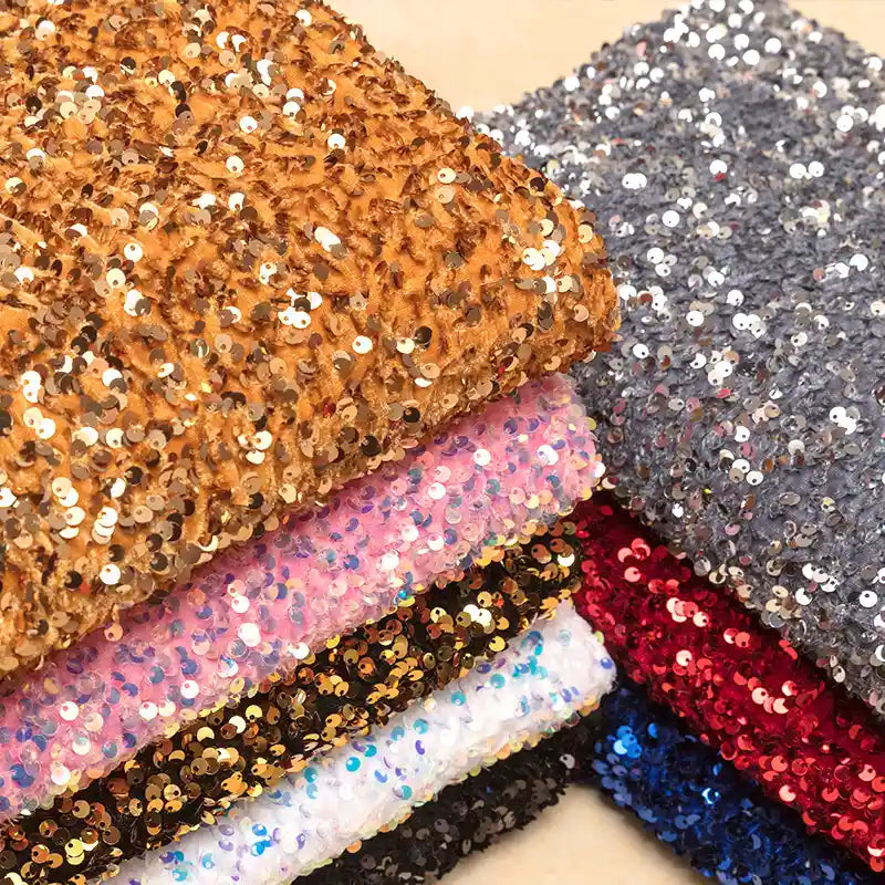 Une pile de Tissu en velours extensible à paillettes au mètre de Longan Craft France dans des tons dorés, argentés, roses, noirs, blancs, rouges et multicolores avec une finition pailletée - parfait pour les vêtements de scène et la mode.