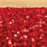 Un gros plan du tissu en velours extensible à paillettes au mètre de Longan Craft France montre des paillettes rouges brillantes sur un tissu étincelant, reflétant la lumière. L'arrière-plan est flou et de couleur crème. Idéal pour les vêtements de scène et les créations de mode.