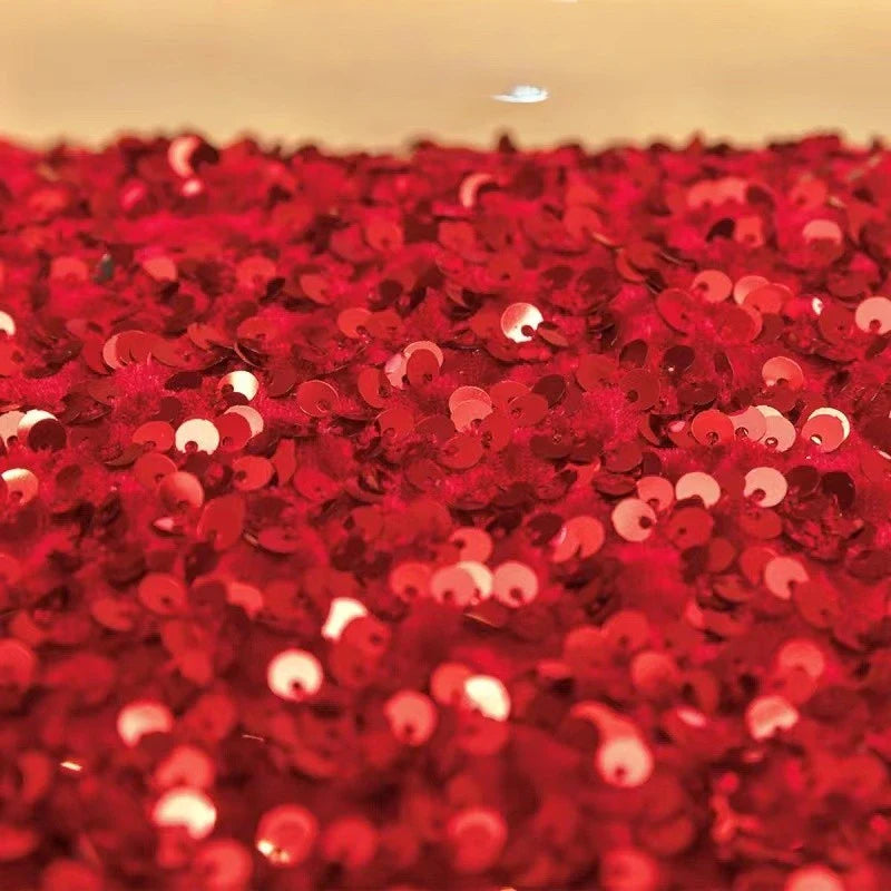 Un gros plan du tissu en velours extensible à paillettes au mètre de Longan Craft France montre des paillettes rouges brillantes sur un tissu étincelant, reflétant la lumière. L'arrière-plan est flou et de couleur crème. Idéal pour les vêtements de scène et les créations de mode.