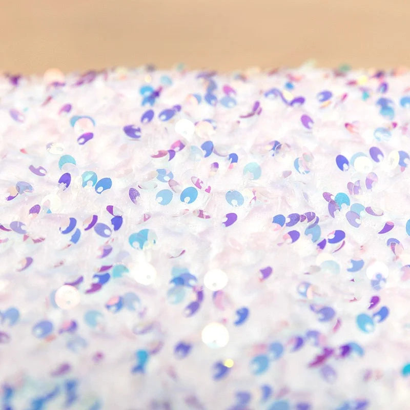 Gros plan sur Tissu en velours extensible à paillettes au mètre de Longan Craft France, montrant un tissu texturé blanc avec des paillettes iridescentes violettes, bleues et roses créant un motif étincelant et coloré idéal pour les vêtements de scène et la mode.