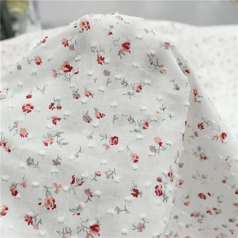 Le "Tissu plumetis rouge à motifs floraux au mètre" de Longan Craft France présente une texture pointillée vintage avec de petits motifs floraux rouges, roses et violets, parfaits pour des créations élégantes.