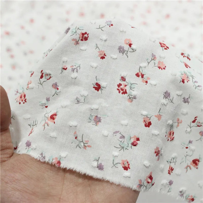 Une main tient un morceau de tissu plumetis rouge à motifs floraux au mètre de Longan Craft France, avec des points vintage en relief et de minuscules fleurs rouges, roses et violettes aux tiges vertes. D'autres pièces de ce charmant tissu rétro apparaissent à l'arrière-plan.