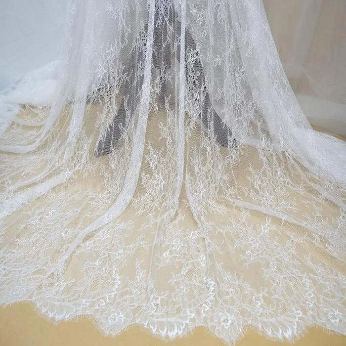 Dentelle Chantilly blanche au mètre de Longan Craft France : tissu dentelle français floral à bord festonné, parfait pour robes de mariée DIY, drapé sur fond beige avec motifs floraux délicats et détails raffinés.