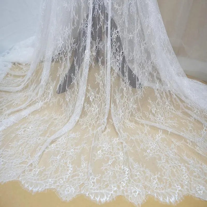 Dentelle Chantilly blanche au mètre de Longan Craft France : tissu dentelle français floral à bord festonné, parfait pour robes de mariée DIY, drapé sur fond beige avec motifs floraux délicats et détails raffinés.