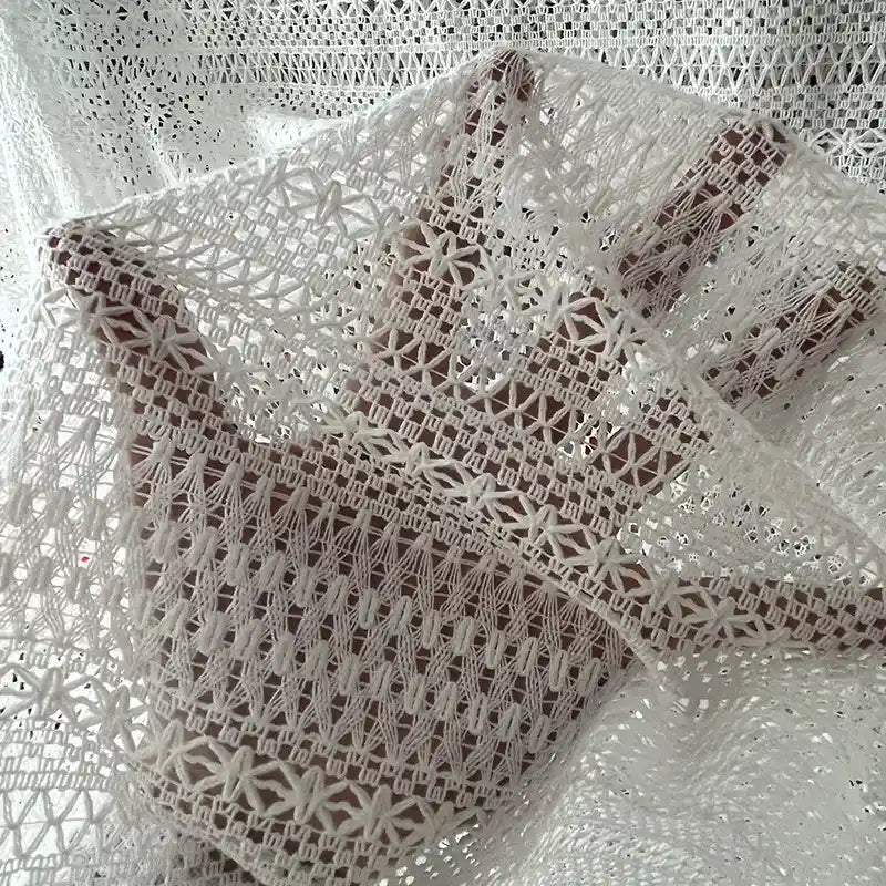 Une main est montrée sous le tissu dentelle en coton blanc au mètre de Longan Craft France, une dentelle jacquard ajourée idéale pour DIY et confection de robes, laissant apparaître la main à travers son motif ajouré semi-transparent.