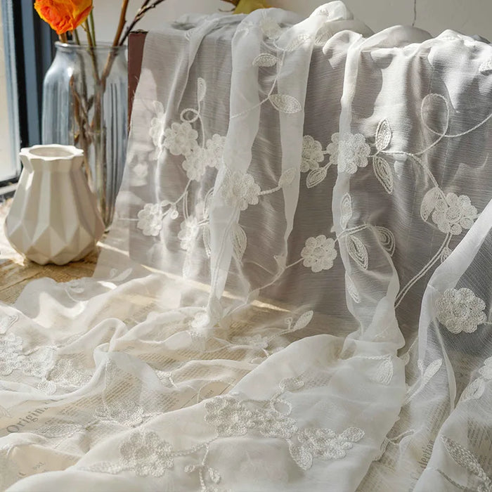 Le Tissu mousseline transparente brodée de fleurs au mètre de Longan Craft France est drapé à côté d'un vase blanc texturé et d'un vase en verre avec des fleurs orange, baignés par la lumière du soleil à travers la fenêtre.