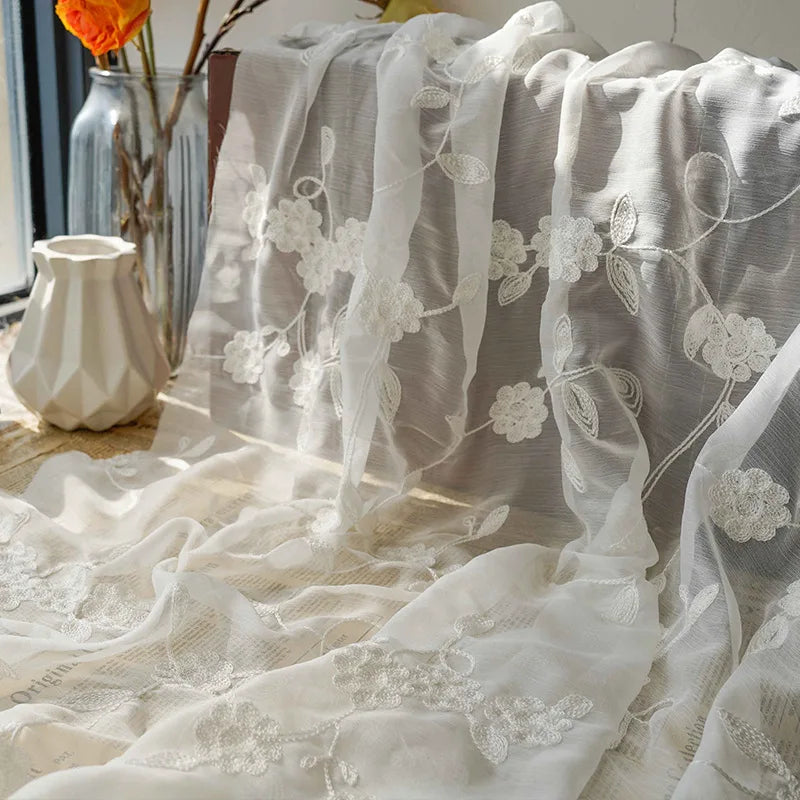 Le Tissu mousseline transparente brodée de fleurs au mètre de Longan Craft France est drapé à côté d'un vase blanc texturé et d'un vase en verre avec des fleurs orange, baignés par la lumière du soleil à travers la fenêtre.
