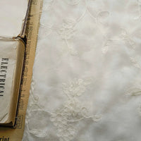 Gros plan sur le tissu mousseline transparent brodé de fleurs au mètre de Longan Craft France, idéal pour créer des vêtements élégants et légers, présenté à côté d'un vieux livre et d'un journal jauni avec un texte partiel.