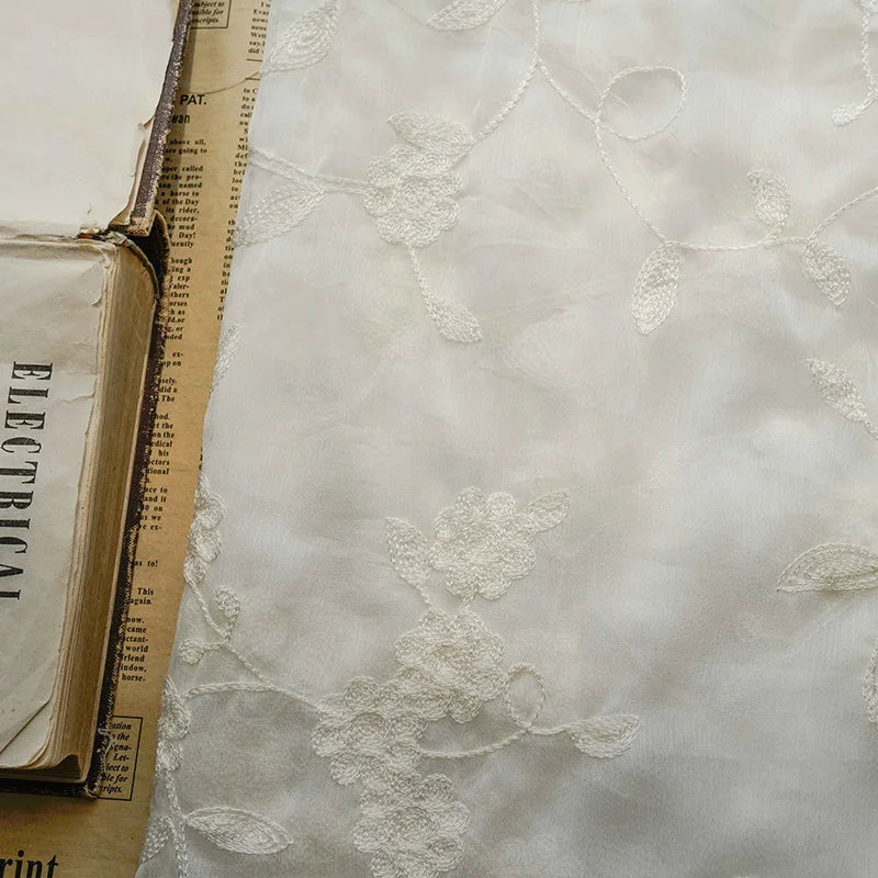 Gros plan sur le tissu mousseline transparent brodé de fleurs au mètre de Longan Craft France, idéal pour créer des vêtements élégants et légers, présenté à côté d'un vieux livre et d'un journal jauni avec un texte partiel.
