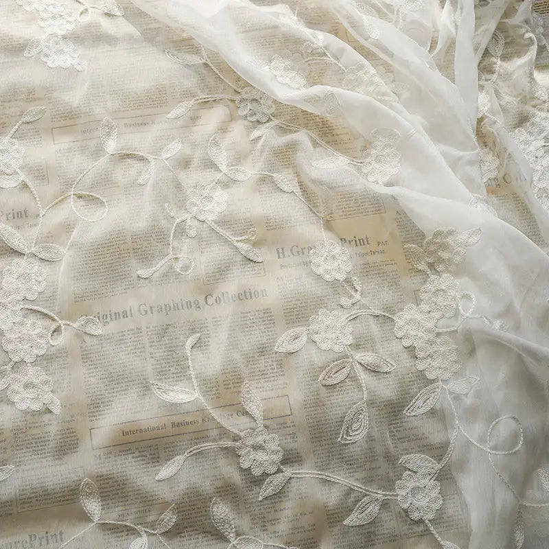 Le Tissu mousseline transparente brodée de fleurs au mètre de Longan Craft France présente une broderie florale transparente superposée à un journal imprimé, créant un contraste saisissant entre la dentelle délicate et le fond graphique.