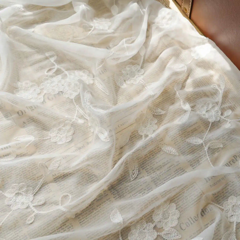 Un morceau de Tissu mousseline transparente brodée de fleurs au mètre de Longan Craft France est drapé sur des pages de journaux, avec une surface en cuir marron visible dans le coin supérieur droit.
