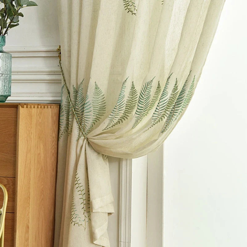 Un rideau de tulle blanc à motifs feuillus de Longan Craft France est attaché par un meuble en bois, révélant partiellement un mur blanc et des moulures décoratives.