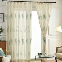 Tissu de rideau en tulle blanc à motifs feuilles Longan Craft France habille une tringle noire, partiellement tiré sur le côté. La pièce présente des murs blancs, un lampadaire, une table d'appoint et un fauteuil avec plaid en fourrure.