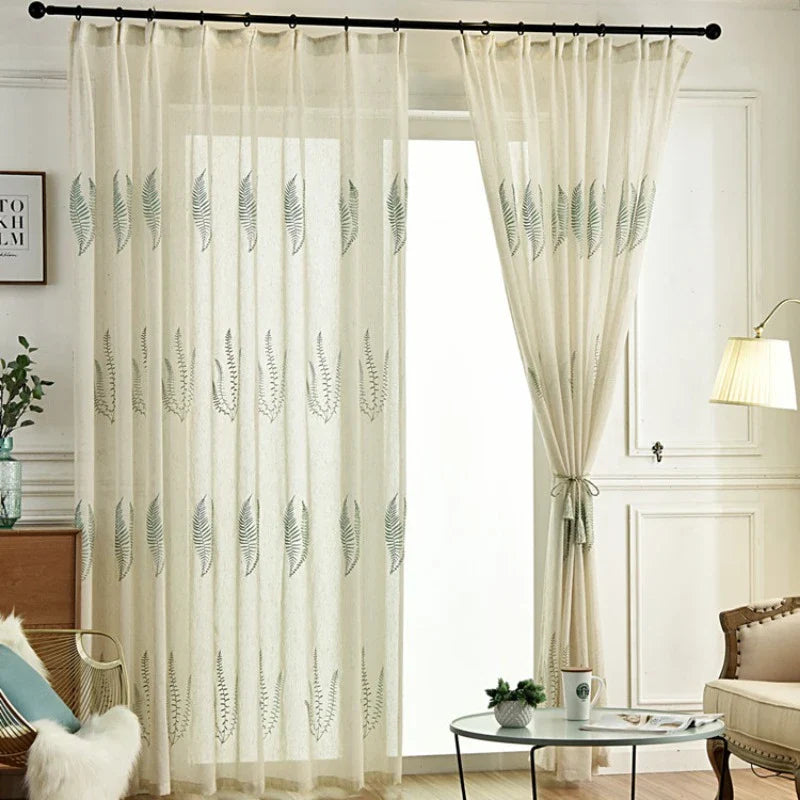 Tissu de rideau en tulle blanc à motifs feuilles Longan Craft France habille une tringle noire, partiellement tiré sur le côté. La pièce présente des murs blancs, un lampadaire, une table d'appoint et un fauteuil avec plaid en fourrure.