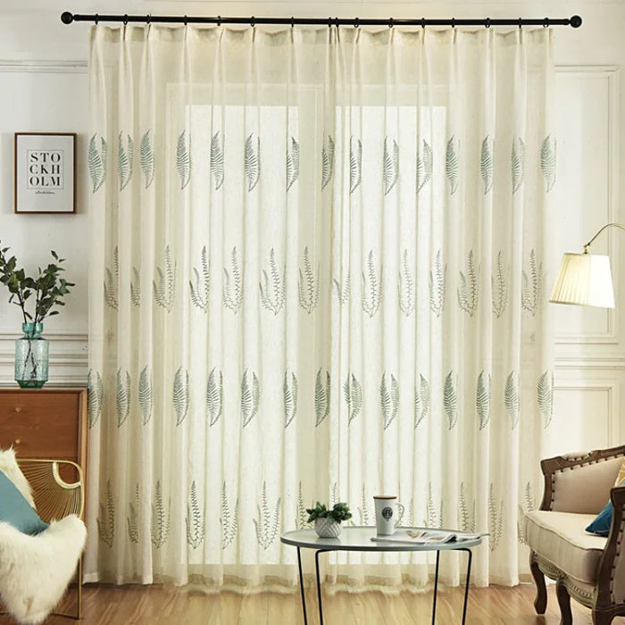 Un salon avec Longan Craft France Tissu de rideau en tulle blanc à motifs feuilles, une table en bois contenant une tasse à café, une chaise en rotin, une plante sur une table d'appoint, un lampadaire et un décor encadré. La lumière du soleil traverse les rideaux botaniques transparents.