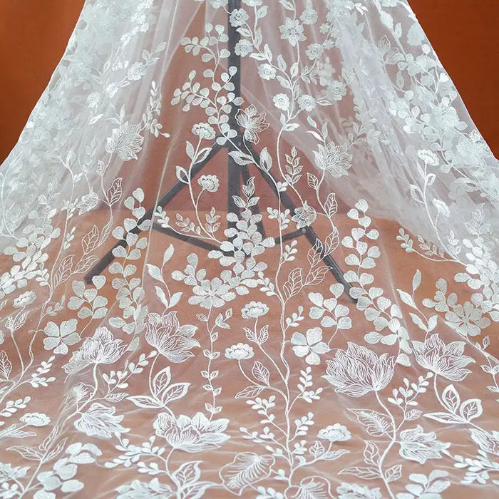 Le Tulle brodé floral blanc au mètre pour couture de robe élégante de Longan Craft France présente des fleurs, des feuilles et des branches brodées en blanc sur du tulle transparent - idéal pour les robes de cérémonie et les projets de couture élégants.