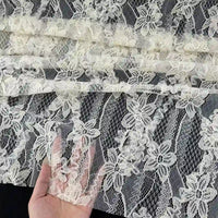 Une main soulève une section de Tissu en dentelle 3D au mètre de Longan Craft France, avec des fleurs et feuilles brodées, présentées sur un fond sombre pour mettre en valeur l'élégante matière dentelle française pour mode printanière et estivale.