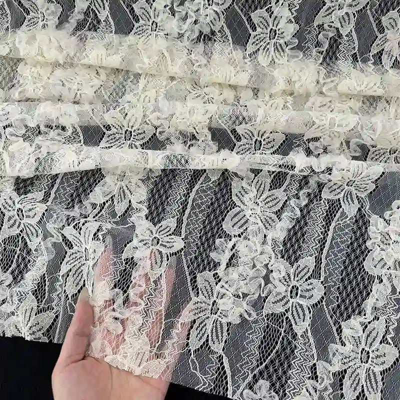 Une main soulève une section de Tissu en dentelle 3D au mètre de Longan Craft France, avec des fleurs et feuilles brodées, présentées sur un fond sombre pour mettre en valeur l'élégante matière dentelle française pour mode printanière et estivale.