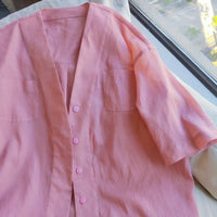 Une chemise rose pâle à manches courtes et à boutons, avec deux poches sur la poitrine, se trouve près d'une fenêtre ensoleillée - une inspiration parfaite pour créer votre propre "Patron de chemise oversize femme à col en V" avec le patron PDF gratuit de Longan Craft France.