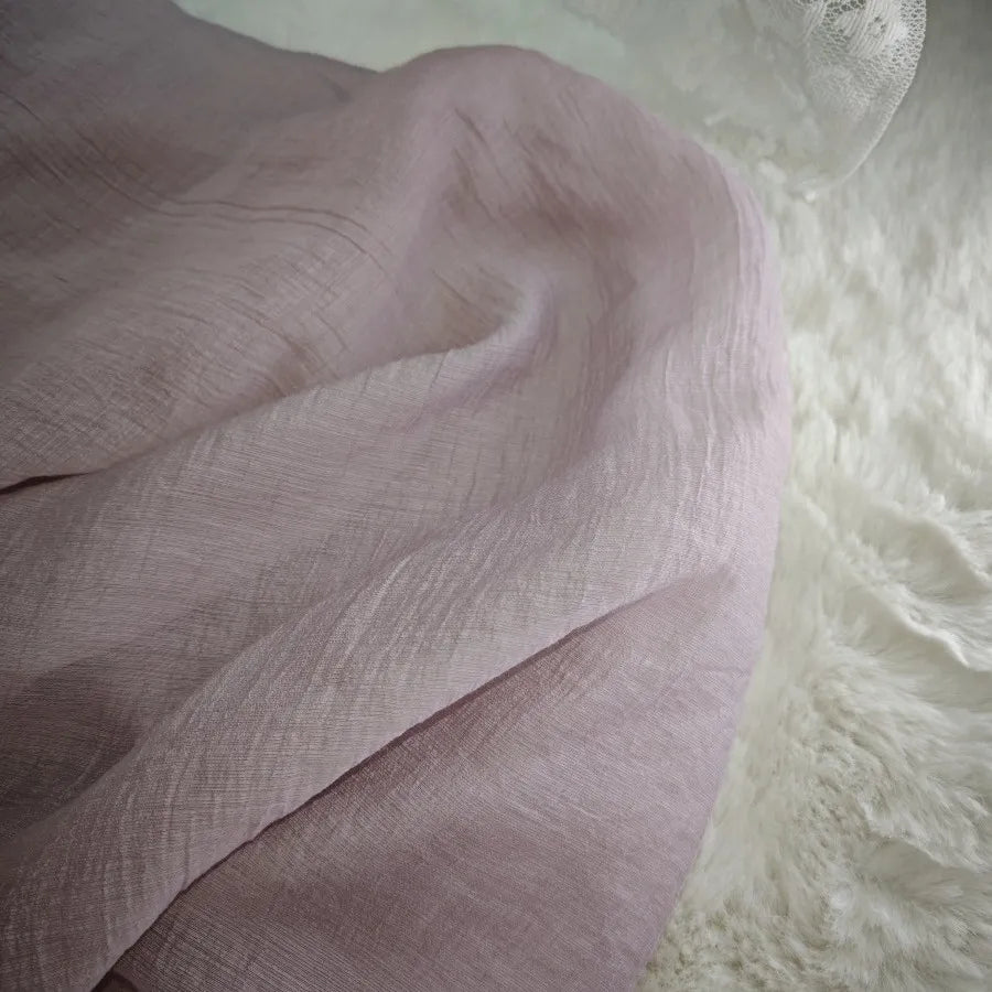 Gros plan sur le tissu lyocell froissé au mètre de Longan Craft France, un textile mauve clair, respirant et durable pour les hauts, drapé sur une surface blanche douce ressemblant à de la fourrure avec des détails de dentelle dans le coin supérieur droit.