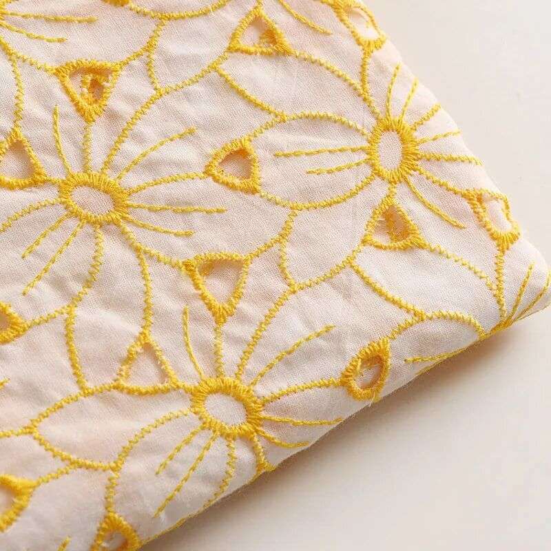 Gros plan sur le Tissu brodé ajouré œillet jaune au mètre de Longan Craft France, un tissu brodé 100 % coton avec des détails floraux jaunes découpés, plié proprement sur une surface légère.