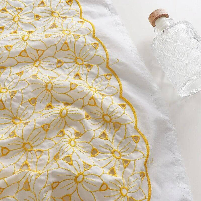 Le tissu brodé ajouré œillet jaune au mètre de Longan Craft France, un tissu 100 % coton présentant une broderie florale jaune complexe, est présenté à côté d'une bouteille en verre transparent avec bouchon en liège sur une surface blanche.