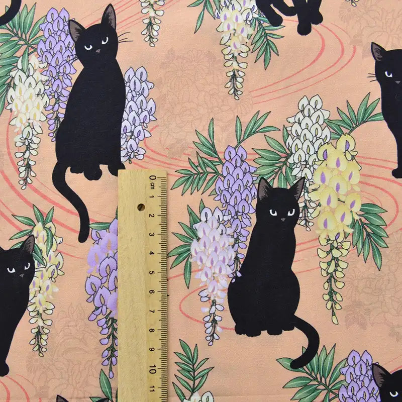 Le Tissu popeline coton imprimé floral chat de Longan Craft France représente des chats noirs avec des glycines violettes et blanches sur des pêches, présentés avec une règle en bois pour l'échelle. Idéal pour le bricolage, le patchwork, les robes, les chemises, ou la décoration intérieure.