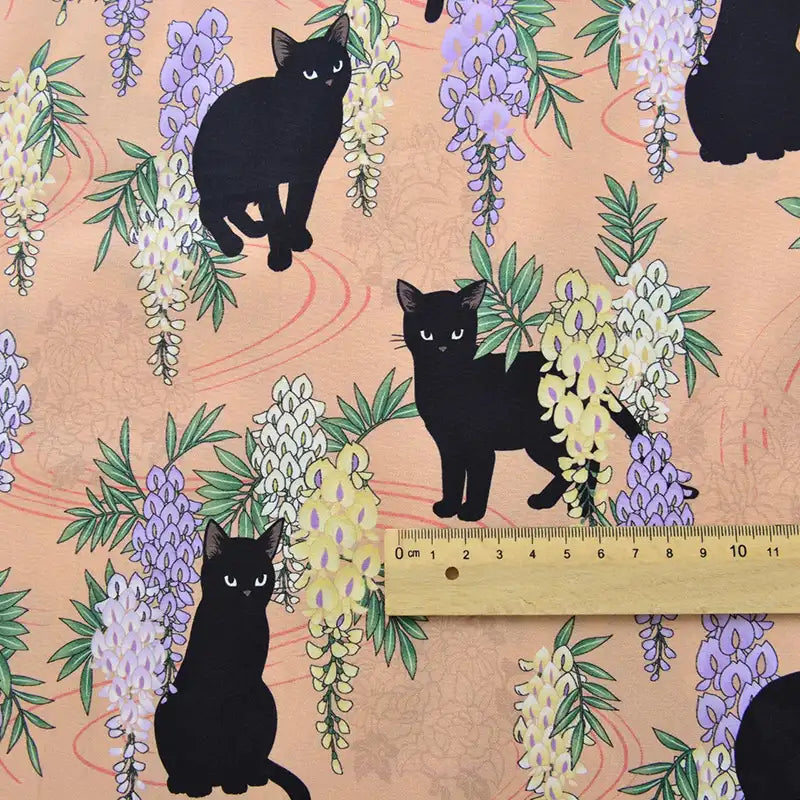 Le Tissu popeline coton imprimé floral chat au mètre de Longan Craft France représente des chats noirs parmi des glycines violettes sur un fond pêche - parfait pour le bricolage, le patchwork, les peignoirs, les chemises et la décoration intérieure.