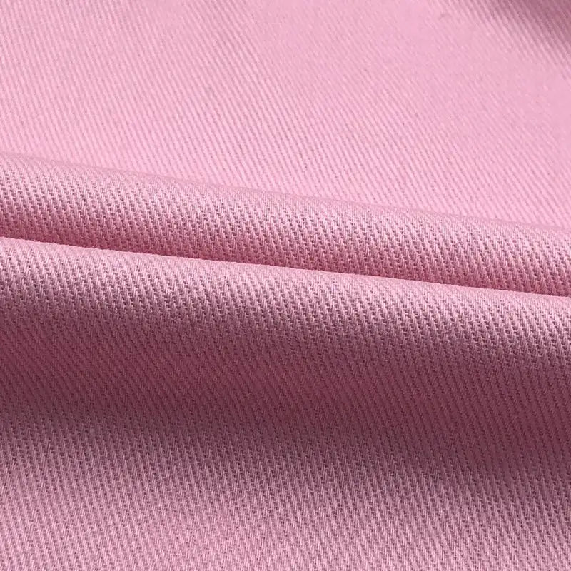 Gros plan sur le Tissu jean coloré 7,5 oz au mètre de Longan Craft France : denim sergé rose pâle, 100 % coton, avec une armure sergée diagonale et une texture douce, présentant un léger pli horizontal sur le tissu.