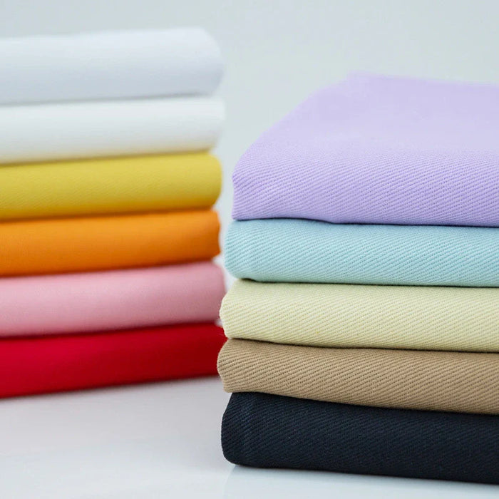Les piles de Tissu jean coloré 7,5 oz au mètre de Longan Craft France - denim sergé multicolore 100 % coton - sont présentées dans des couleurs vibrantes sur une surface blanche réfléchissante, parfaite pour les jeans, les vêtements et la décoration.