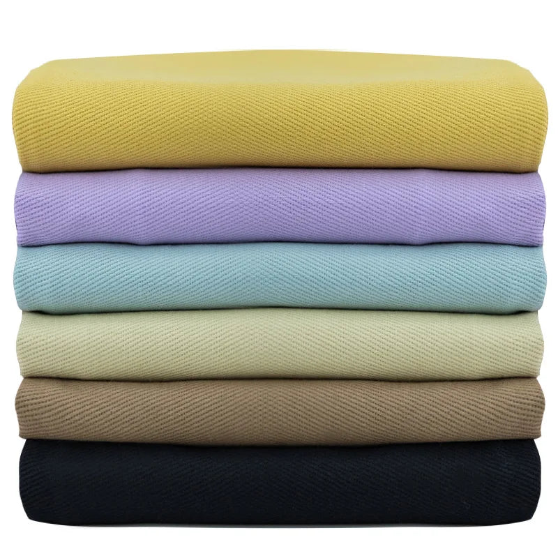 Une pile de six Tissu jean coloré 7,5 oz au mètre par Longan Craft France en jaune, lavande, bleu, vert, tan, et noir est soigneusement arrangée sur un fond blanc. Denim sergé multicolore 100% coton pour jeans colorés.