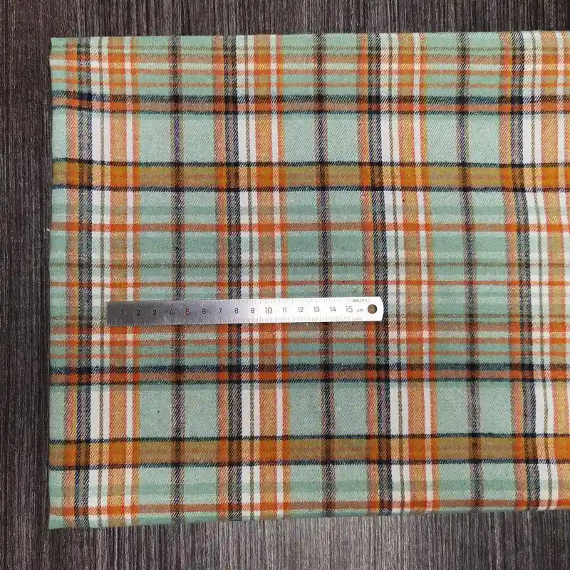 Une règle métallique de 15 cm repose sur le "Tissu en flanelle à carreaux colorés au mètre" de Longon Craft France, une flanelle douce en polyester-coton idéale pour l'automne, présentée en vert, orange, blanc et noir sur une surface texturée gris foncé.