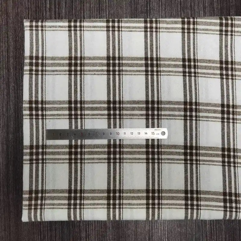 Une règle en métal est posée horizontalement sur un tissu en flanelle à carreaux colorés au mètre de Longon Craft France, montrant son motif à carreaux noirs et bruns, sur une surface texturée foncée. La règle mesure environ 18 cm.