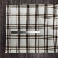 Une règle en métal est posée horizontalement sur un tissu en flanelle à carreaux colorés au mètre de Longon Craft France, montrant son motif à carreaux noirs et bruns, sur une surface texturée foncée. La règle mesure environ 18 cm.