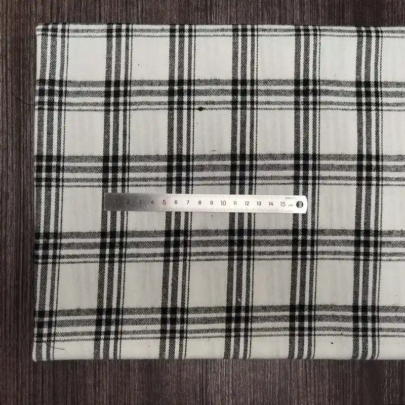 Une règle argentée de 15 cm repose sur une surface blanche recouverte de tissu en flanelle à carreaux colorés de Longon Craft France, un doux mélange de polyester et de coton parfait pour l'automne. L'arrière-plan est en bois foncé.