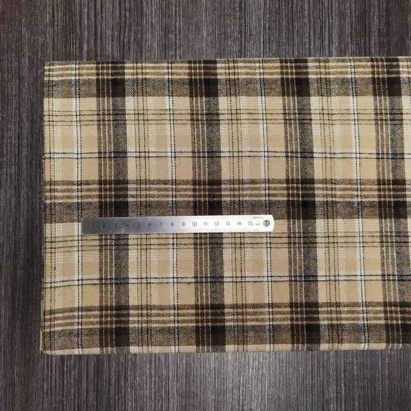 Une règle en métal est posée sur le tissu en flanelle à carreaux colorés au mètre de Longon Craft France, un tissu douillet en polyester-coton parfait pour l'automne, présenté sur une surface en bois sombre. La règle indique les mesures métriques.
