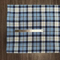 Une règle métallique de 15 cm repose sur le "Tissu en flanelle à carreaux colorés au mètre" de Longon Craft France, un tissu écossais douillet en polyester et coton, présenté sur une surface texturée brune.