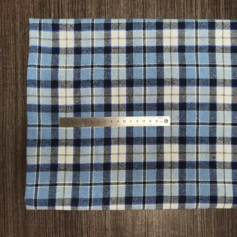 Une règle métallique de 15 cm repose sur le "Tissu en flanelle à carreaux colorés au mètre" de Longon Craft France, un tissu écossais douillet en polyester et coton, présenté sur une surface texturée brune.