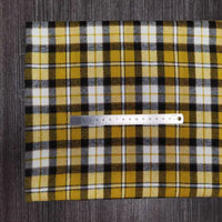 Une règle métallique de 15 cm est posée sur le tissu en flanelle à carreaux colorés au mètre de Longon Craft France, une flanelle en polyester-coton pour l'automne, présentée en jaune, noir et blanc sur une surface texturée gris foncé.