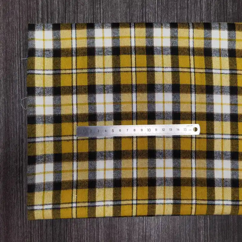 Une règle métallique de 15 cm est posée sur le tissu en flanelle à carreaux colorés au mètre de Longon Craft France, une flanelle en polyester-coton pour l'automne, présentée en jaune, noir et blanc sur une surface texturée gris foncé.