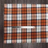 Une règle métallique de 15 cm est placée sur le tissu Longon Craft France Tissu en flanelle à carreaux colorés au mètre, un tissu confortable en polyester-coton pour l'automne, présenté en orange, noir, blanc et bleu sur une surface texturée foncée.