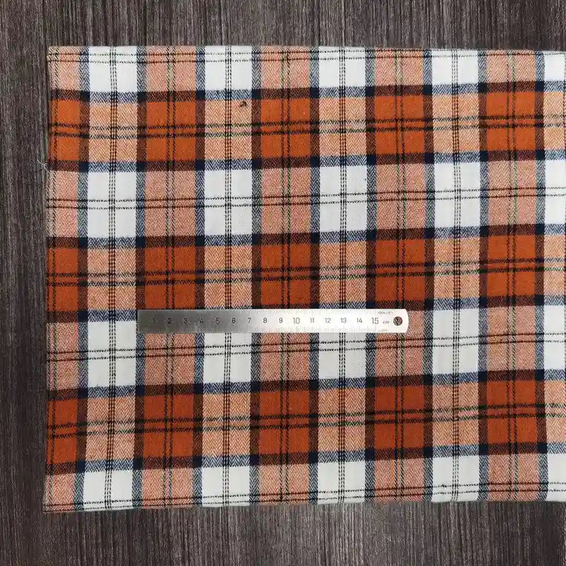 Une règle métallique de 15 cm est placée sur le tissu Longon Craft France Tissu en flanelle à carreaux colorés au mètre, un tissu confortable en polyester-coton pour l'automne, présenté en orange, noir, blanc et bleu sur une surface texturée foncée.
